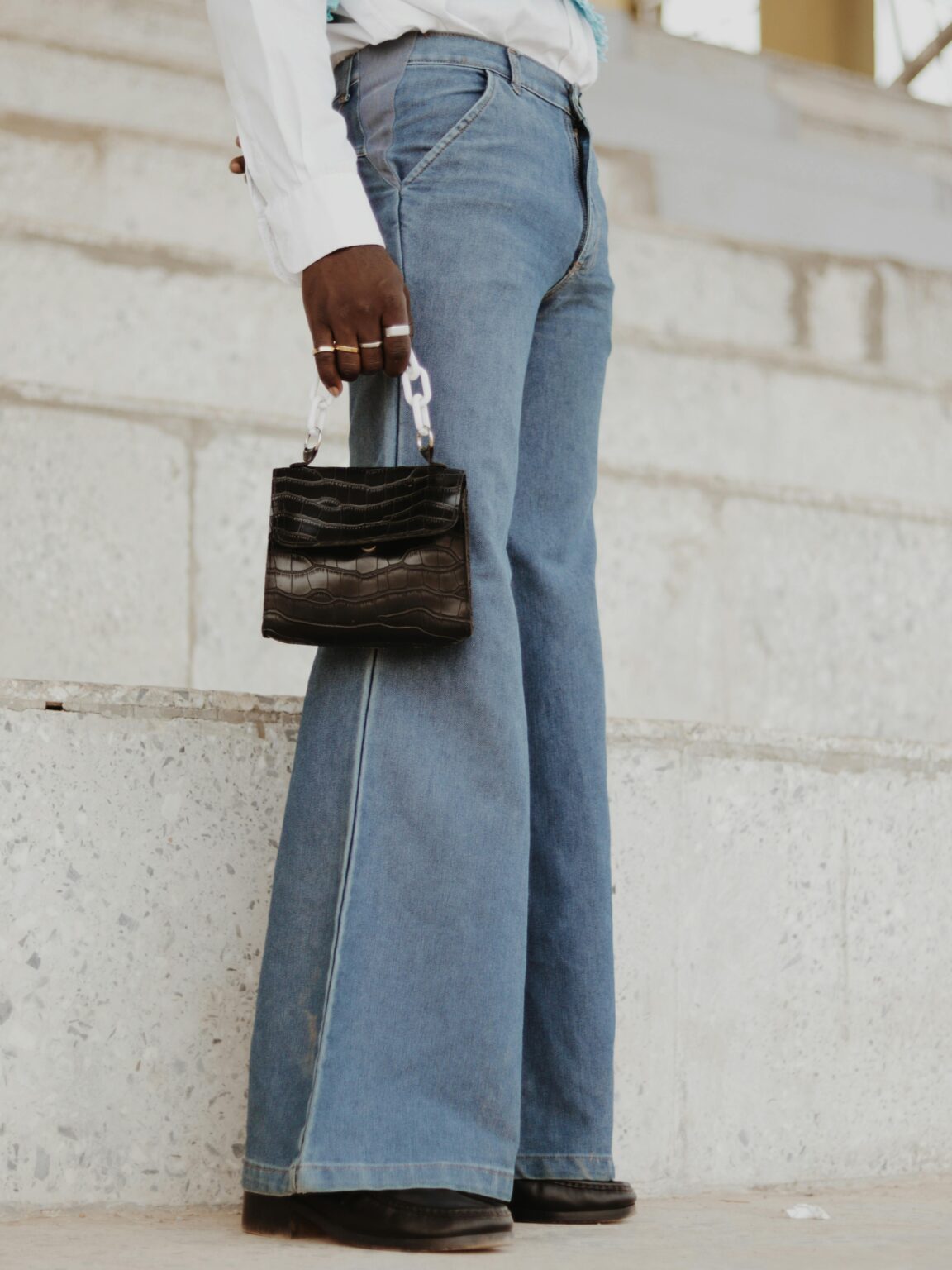 Fashionable adult holding a mini black handbag while wearing bell-bottom jeans outdoors.