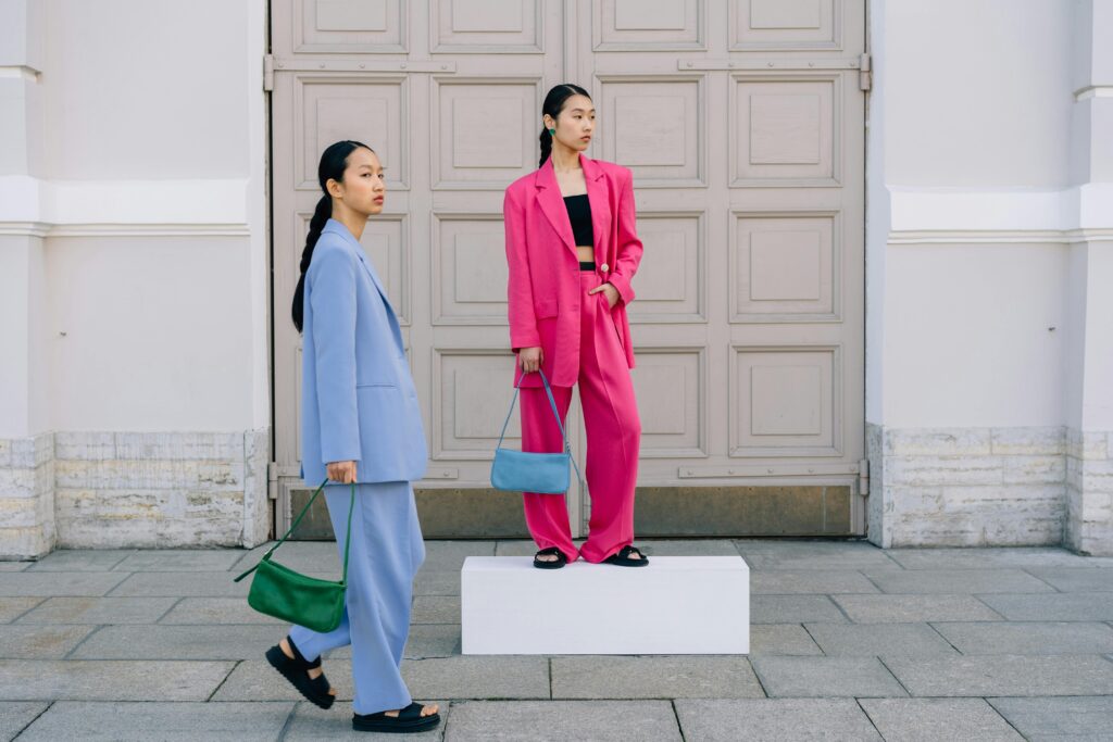 Two fashionable women in vibrant blazers pose with stylish handbags outside a vintage building.